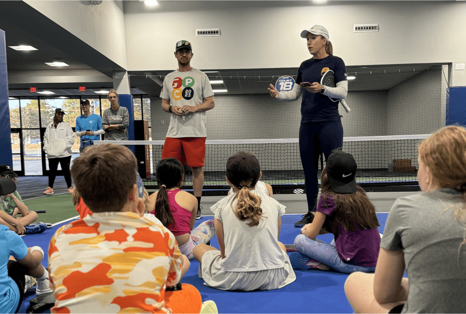 National Junior Pickleball - Group of children sit on a court, listening to two instructors